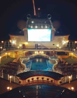 A luxury cruise ship deck featuring a pool surrounded by lounge chairs and warm ambient lighting. A large screen overlooks the area, showing an image against a dark night sky.