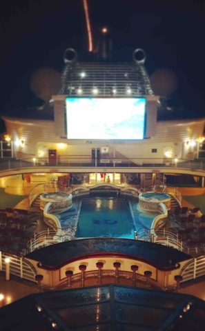 A luxury cruise ship deck featuring a pool surrounded by lounge chairs and warm ambient lighting. A large screen overlooks the area, showing an image against a dark night sky.