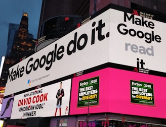 A bustling urban setting with large digital billboards displaying colorful advertisements. Prominent ads include 'Make Google do it' and 'American Idol Winner David Cook'. Bright neon colors and giant screens dominate the scene, hinting at a lively nightlife area.