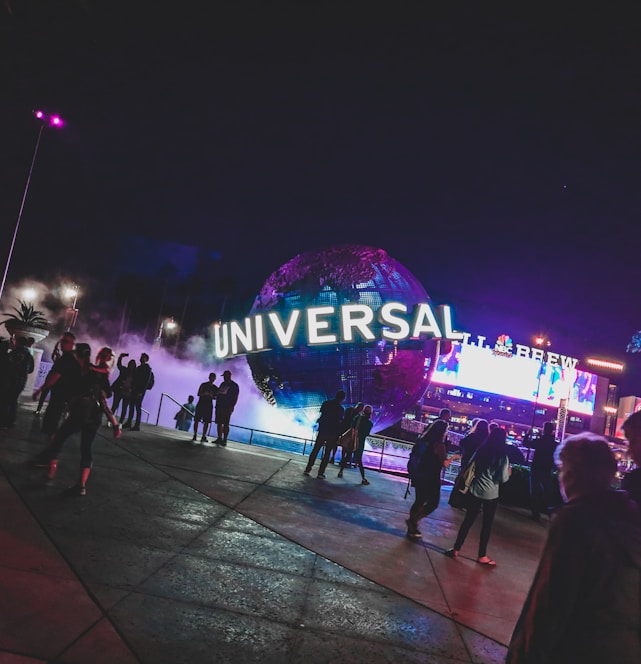 A large illuminated globe with the word 'Universal' is prominent at the center, surrounded by a misty, atmospheric effect. People are walking around the area, with some standing and looking at the globe. In the background, there are bright electronic billboards and palm trees, contributing to a lively and bustling environment.