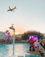 A vibrant outdoor party at a poolside venue with a large crowd gathered around a stage. A plane is flying overhead in the clear sky, and a creative water feature in the form of a giant floating tap is pouring water into the pool. Bright lights and a colorful digital screen on the stage add to the energetic atmosphere.