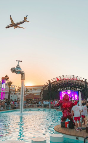 A vibrant outdoor party at a poolside venue with a large crowd gathered around a stage. A plane is flying overhead in the clear sky, and a creative water feature in the form of a giant floating tap is pouring water into the pool. Bright lights and a colorful digital screen on the stage add to the energetic atmosphere.