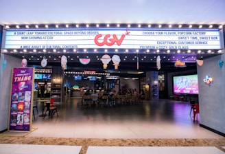 A brightly lit cinema entrance with a marquee displaying various messages. There is a large neon logo above the entrance. Inside, customers are seated at a ticket area, and there are large screens displaying movie-related content. Several decorations hang from the ceiling, adding a festive touch.