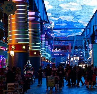 A vibrant indoor complex with illuminated columns and a large digital ceiling display showing a sky pattern. The area is bustling with people, possibly visiting an amusement park or an entertainment venue. There are colorful lights and signage directing to different attractions.