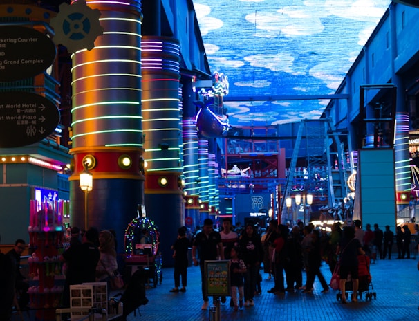 A vibrant indoor complex with illuminated columns and a large digital ceiling display showing a sky pattern. The area is bustling with people, possibly visiting an amusement park or an entertainment venue. There are colorful lights and signage directing to different attractions.