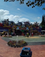 A vibrant amusement park entrance featuring colorful signage and cartoon characters. Visitors gather around a central plaza with landscaped greenery and a small fountain. The area is surrounded by tall trees and lively decorations.