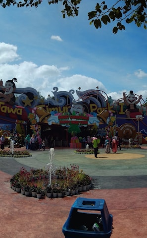 A vibrant amusement park entrance featuring colorful signage and cartoon characters. Visitors gather around a central plaza with landscaped greenery and a small fountain. The area is surrounded by tall trees and lively decorations.