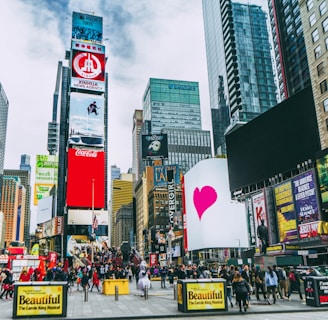 A bustling urban scene with towering skyscrapers and numerous digital billboards showcasing advertisements and entertainment content. A large crowd of people is visible, indicating a busy environment. Bright colors and diverse signage add to the vibrant atmosphere.