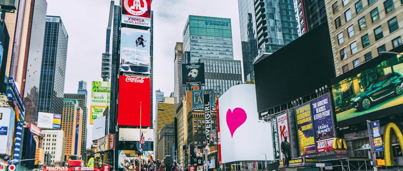 A bustling urban scene with towering skyscrapers and numerous digital billboards showcasing advertisements and entertainment content. A large crowd of people is visible, indicating a busy environment. Bright colors and diverse signage add to the vibrant atmosphere.