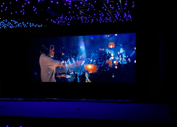 A dimly lit space with a large screen displaying a vibrant scene. The ceiling is adorned with numerous blue lights creating a starry effect. The screen depicts a busy, colorful atmosphere with people, glowing lights, and dynamic motion.
