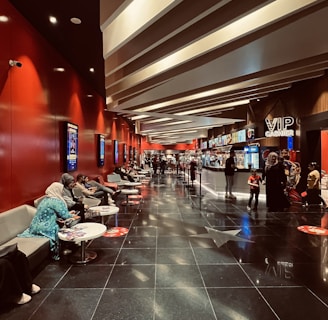 A modern cinema lobby with a sleek design featuring red walls and illuminated ceilings. People are seated on gray couches along the left side, engaging in conversations or using their phones. Posters line the walls, and the floor is polished black with reflective surfaces. Towards the back, there is a counter with a 'VIP Cashier' sign, and a few people are waiting in line.