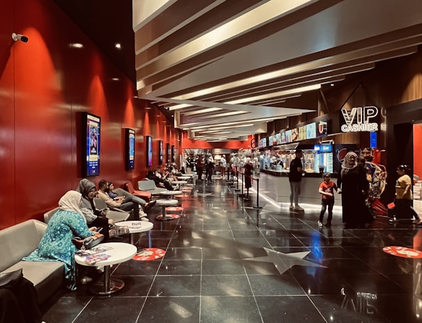 A modern cinema lobby with a sleek design featuring red walls and illuminated ceilings. People are seated on gray couches along the left side, engaging in conversations or using their phones. Posters line the walls, and the floor is polished black with reflective surfaces. Towards the back, there is a counter with a 'VIP Cashier' sign, and a few people are waiting in line.