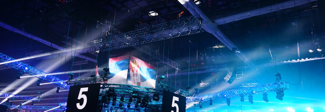 A large indoor arena is equipped with advanced lighting and audio systems. Suspended above the space are numerous digital displays showcasing numbers. The environment is filled with blue illumination and spotlights creating a vibrant atmosphere.