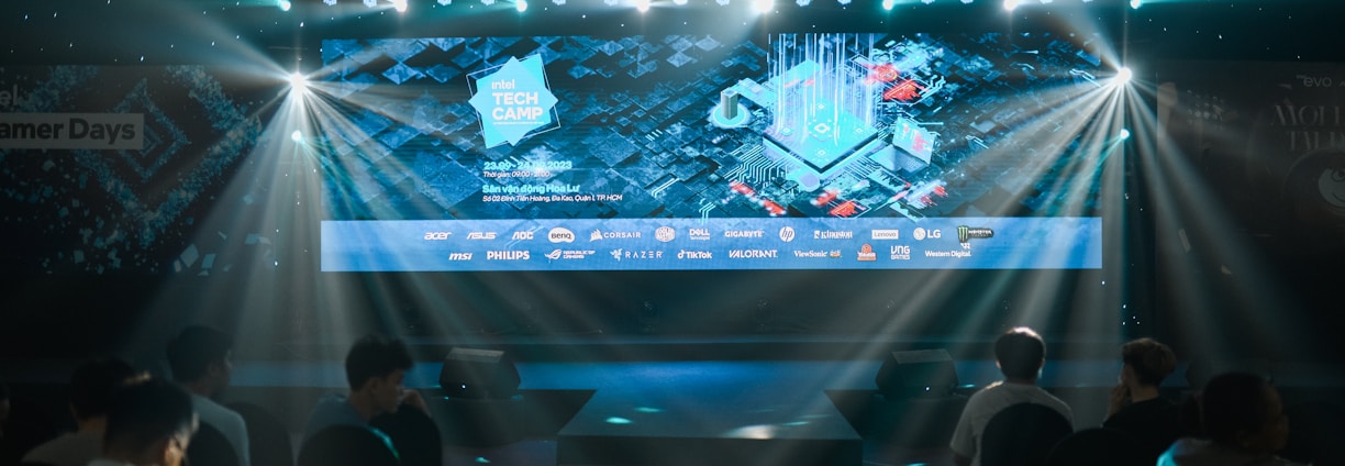 A large illuminated screen displays a tech-themed graphic with logos of various tech companies underneath. The setting appears to be an indoor event or conference with several spotlights beaming down, creating a dynamic atmosphere. Attendees are seated in front, silhouetted against the bright lights.