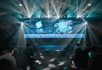 A large illuminated screen displays a tech-themed graphic with logos of various tech companies underneath. The setting appears to be an indoor event or conference with several spotlights beaming down, creating a dynamic atmosphere. Attendees are seated in front, silhouetted against the bright lights.