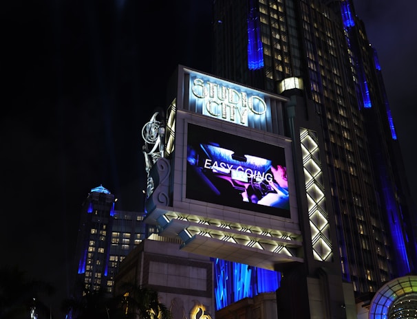 A large, illuminated building at night with the words 'Studio City' prominently displayed. The structure is adorned with bright lights and architectural designs that highlight its grandeur. A large screen above displays an advertisement or video, adding to the lively atmosphere. Surrounding the entrance are palm trees, colorful spotlights, and art deco elements.