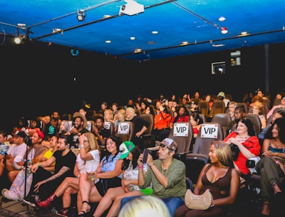 A group of people are seated in a theater, attentively facing the stage. Some seats are marked as 'VIP' in the middle row, indicating a special section. The audience appears diverse, with individuals wearing casual attire. The theater has a blue ceiling with lights, creating a vibrant atmosphere.