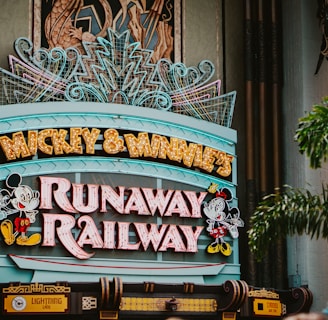 A colorful sign featuring playful typography and animated figures associated with a popular entertainment franchise. The sign is adorned with neon lights and decorative elements, set against a backdrop of bright red pillars and greenery.