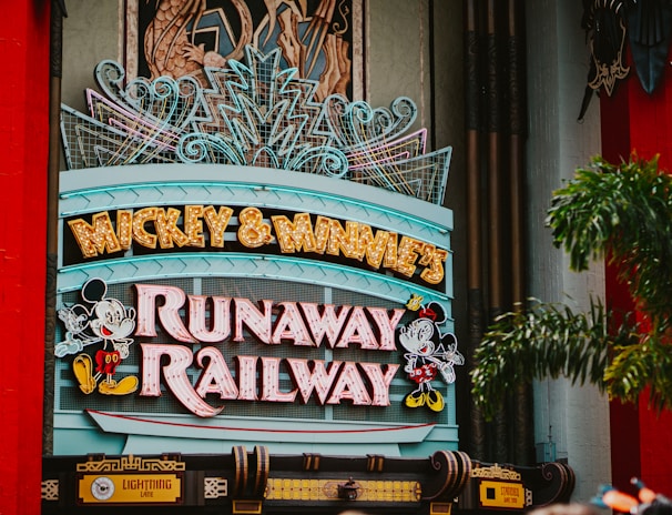 A colorful sign featuring playful typography and animated figures associated with a popular entertainment franchise. The sign is adorned with neon lights and decorative elements, set against a backdrop of bright red pillars and greenery.