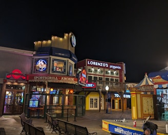 A vibrant night scene showcasing a boardwalk area with various attractions and eateries. Illuminated signs, including advertisements for a pizzeria and a sports grill arcade, are visible. Several benches are positioned along the wooden planks. To the right, a colorful booth and a stage with 'smartwater' signage are featured.