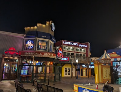 A vibrant night scene showcasing a boardwalk area with various attractions and eateries. Illuminated signs, including advertisements for a pizzeria and a sports grill arcade, are visible. Several benches are positioned along the wooden planks. To the right, a colorful booth and a stage with 'smartwater' signage are featured.