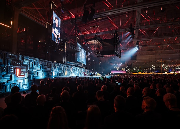 A large indoor event with a stage featuring bright screens and spotlights, attended by a substantial crowd. The venue has a high ceiling with industrial beams and red lighting accents. The atmosphere resembles a concert or a conference with a focus on visual effects.